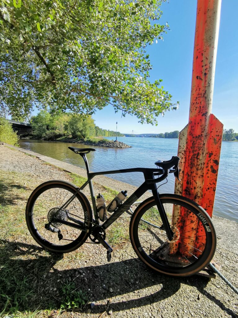 Ein Fahrrad lehnt an einem Mast. Im Hintergrund fließt die Wied von links in den Rhein.